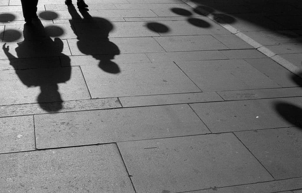 I noticed some interesting rounded sidewalk shadows from lamps hung across the street. But there were a lot of people walking this way, and they often covered up the shadows. It took a bit of waiting until an opening appeared with fewer people. Olympus OM-1, F.Zuiko Auto-S 50 mm f/1.8, Ilford PAN 400